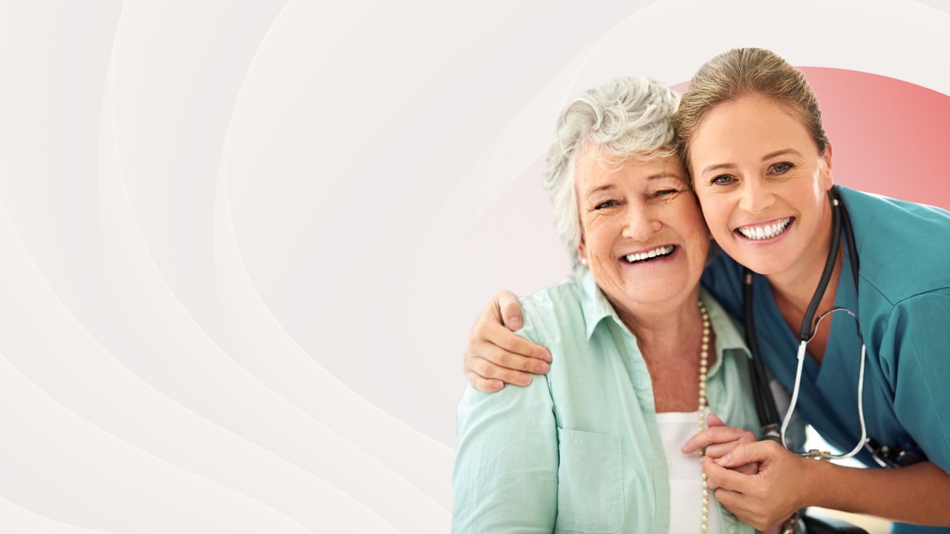 A smiling healthcare worker in blue scrubs with a stethoscope around her neck holds the hand of a smiling older person.