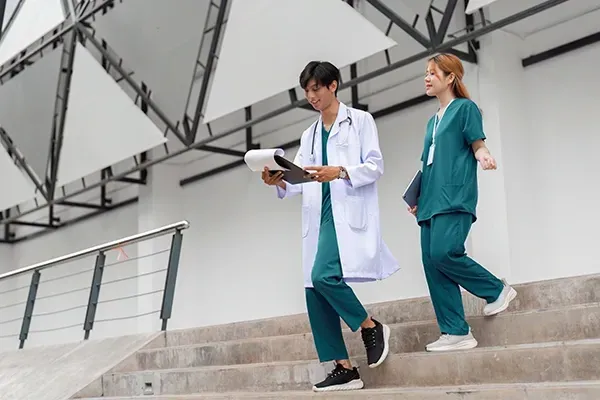A doctor in a white coat and a colleague in green scrubs walk together across a white, tiled hallway.