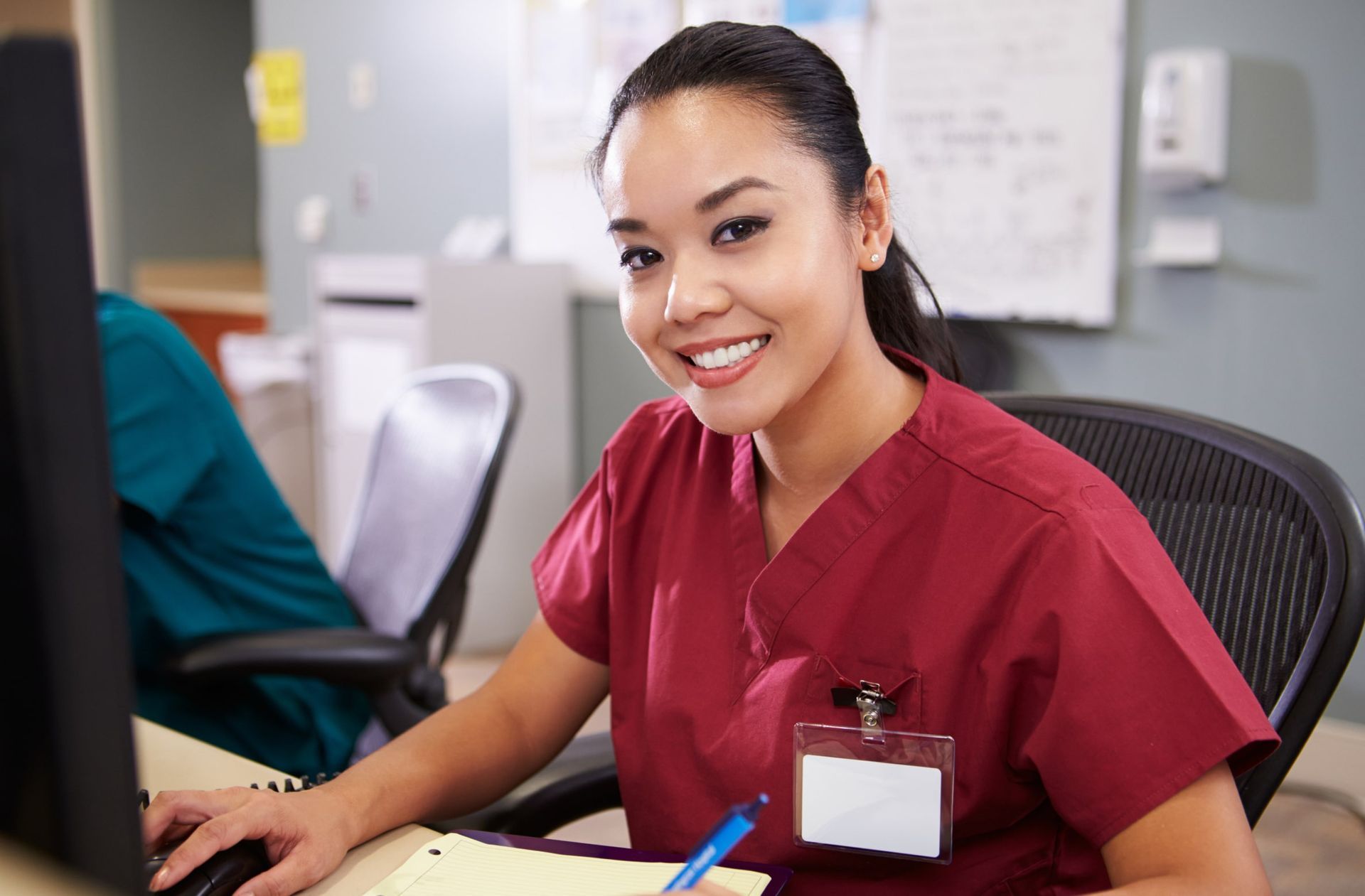 A smiling healthcare worker in maroon scrubs sits at a computer desk, writing on a notepad in a clinical office setting.
