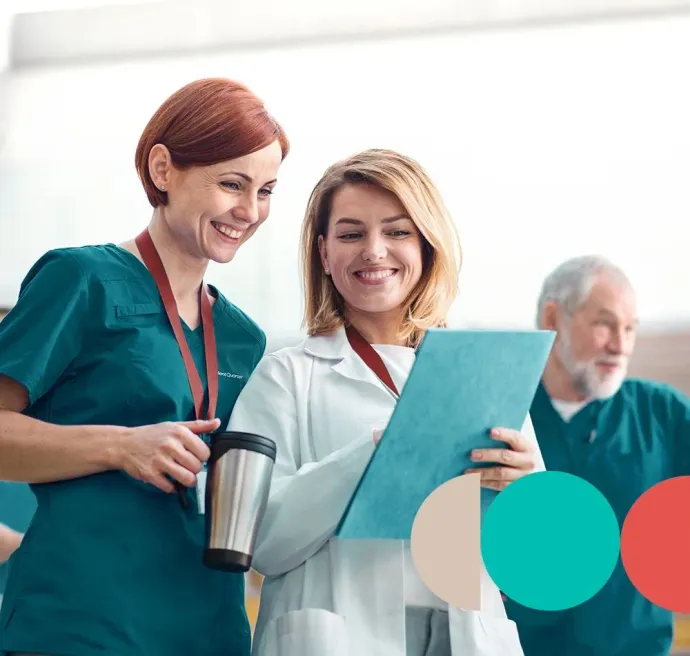 Two healthcare professionals in uniforms review notes on a clipboard and smile, with a colleague visible in the background.