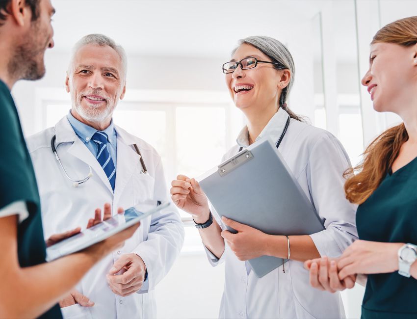 Four medical professionals in scrubs and white coats standing in a bright office, laughing while discussing a tablet.