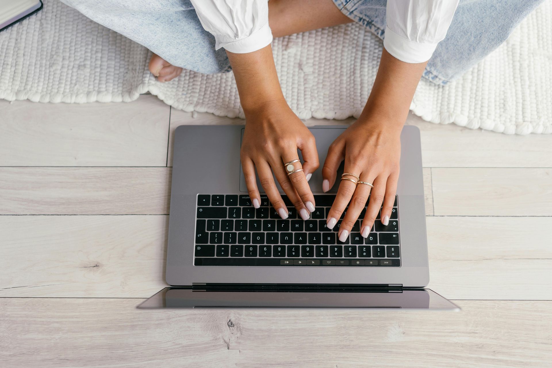 Hands typing on a silver laptop while sitting on a light-colored wooden floor with a white rug.