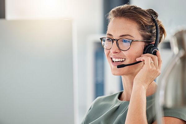 Professional wearing a headset and glasses, smiling while looking at a computer screen in an office.