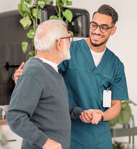 A medical professional in teal scrubs smiling while supporting an elderly person in a grey sweater.