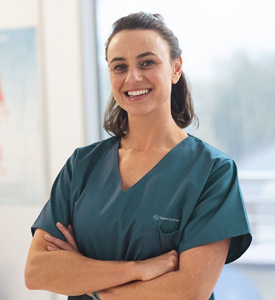 A smiling healthcare professional in a teal scrub top with arms crossed, standing indoors near a window.