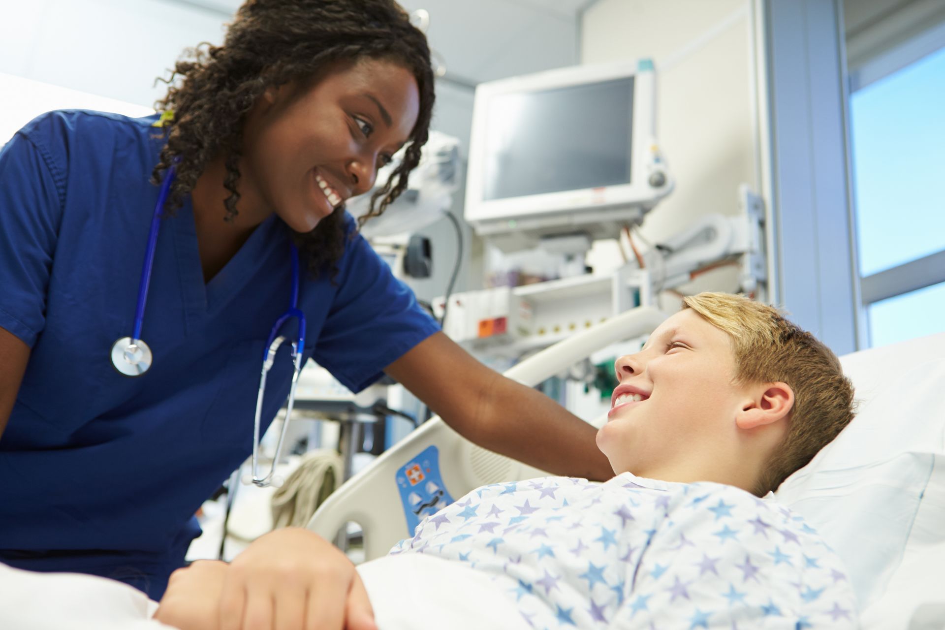 A nurse in blue scrubs smiles while checking on a young patient lying in a hospital bed with medical equipment nearby.