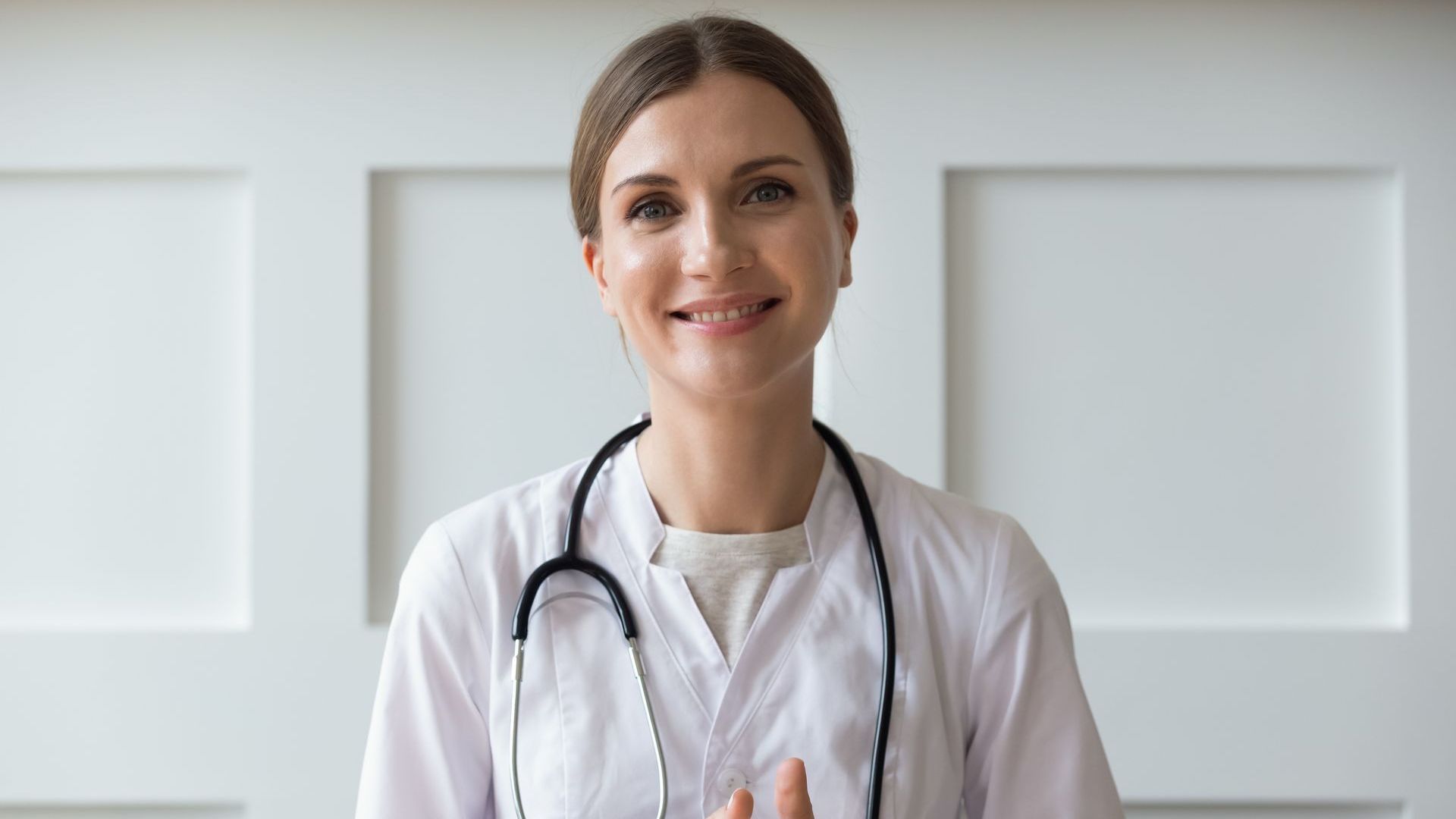 A smiling healthcare professional in a white coat with a stethoscope around their neck, standing against a white wall.