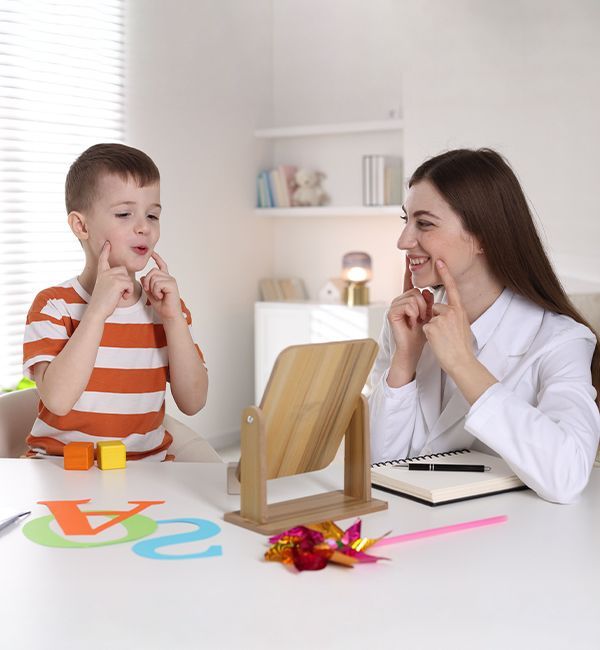 A speech therapist and a child sit at a table using a mirror to practice mouth movements during a therapy session.