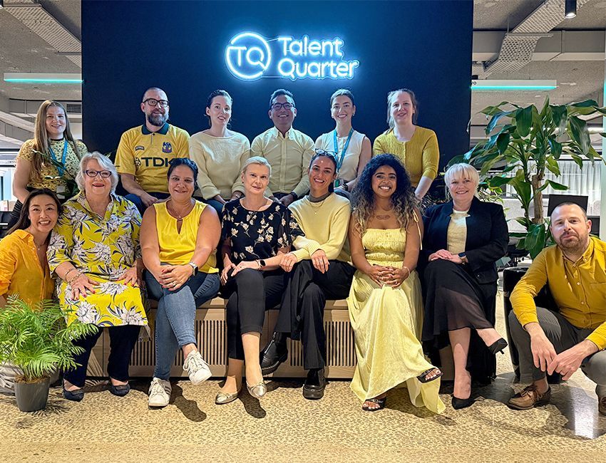 Group of 14 people smiling for a photo in front of a Talent Quarter logo wall, in a modern, plant-filled office setting.