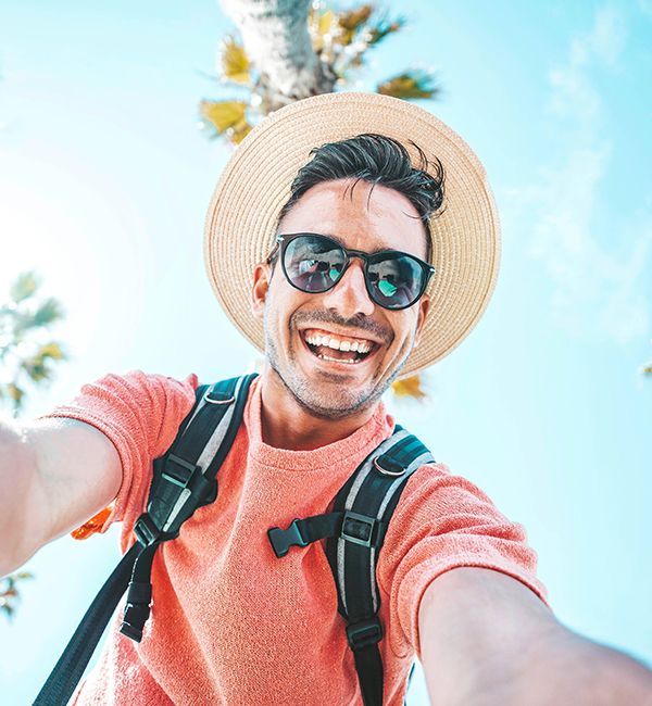 A smiling person wears sunglasses and a straw hat while taking a selfie under bright sky and palm trees.