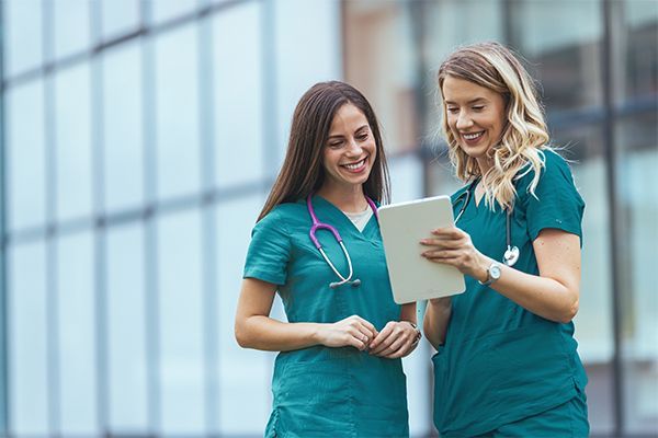 Two healthcare workers in green scrubs stand outside a glass building, smiling while looking at a tablet together.