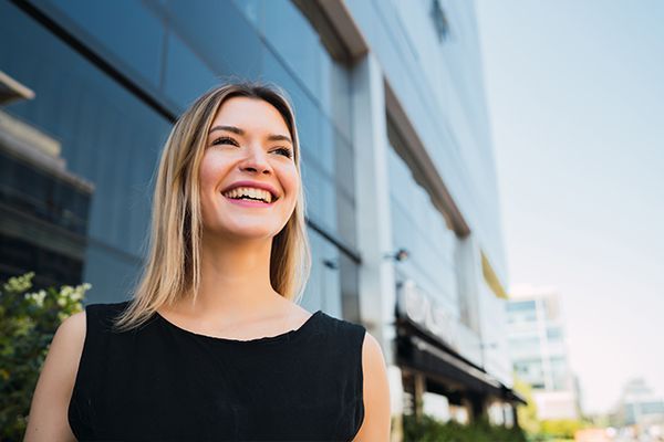 A person with shoulder-length blonde hair smiles while standing outside a modern, glass-paneled building.