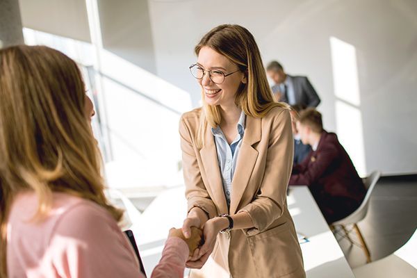 Two people in business attire shaking hands in a bright, modern office with colleagues working in the background.