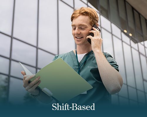 A person in green scrubs holds a folder and speaks on a mobile phone outside a glass building with the text 