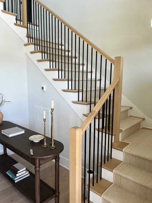 Staircase with black balusters, light wood handrail, and beige carpeted steps beside a hallway table.
