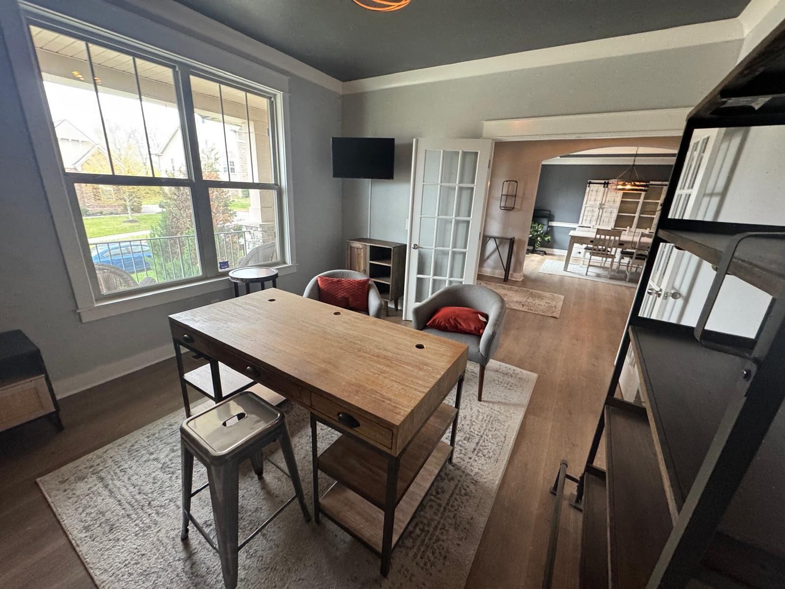 Home office with wooden desk, two stools, large window, wall-mounted TV, and bookshelf leading to a dining area