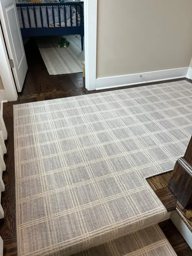 Light gray patterned rug in a hallway, with a doorway leading to a room and wooden floor beyond