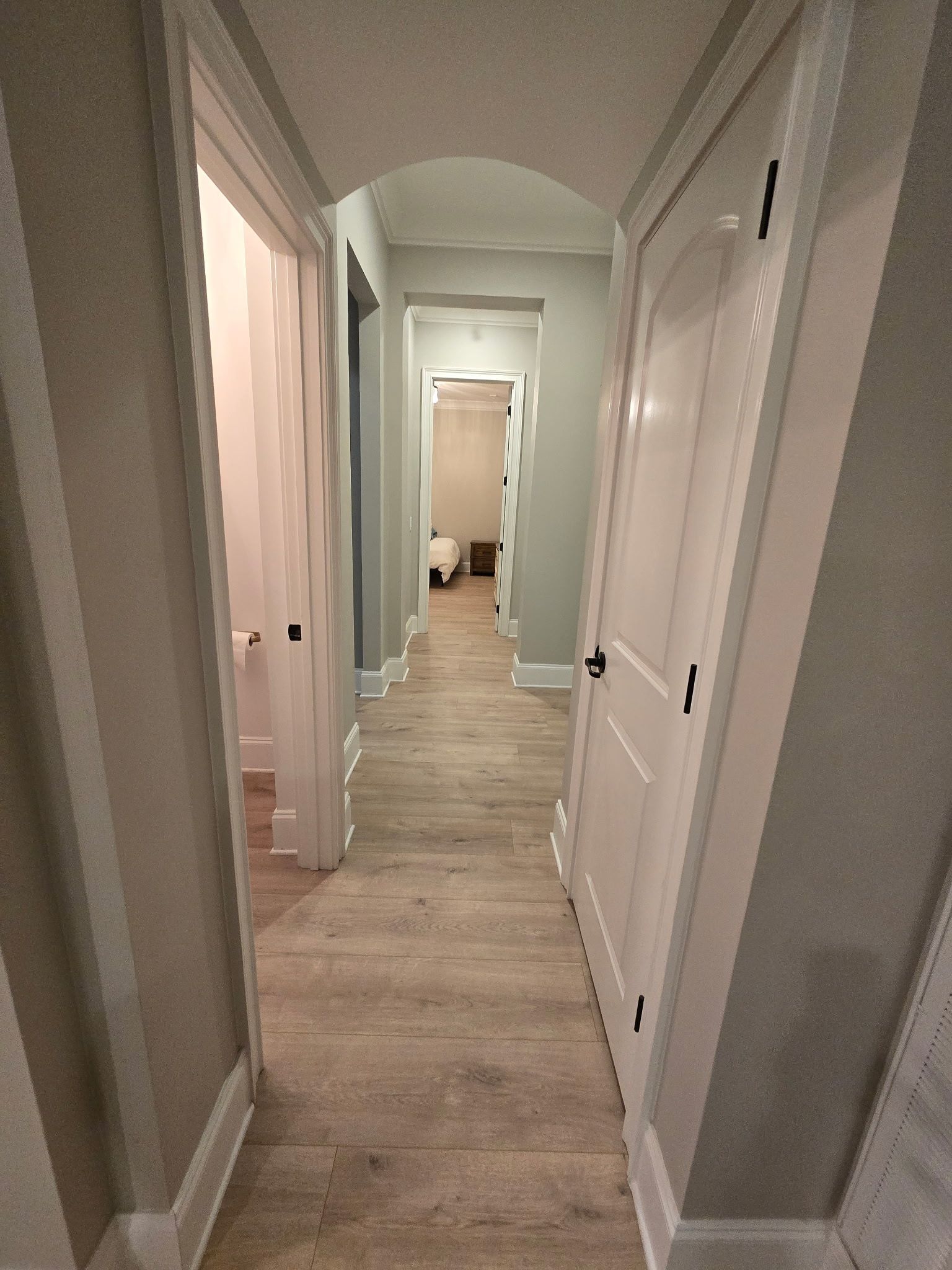 Long hallway with white doors, light wood floor, and arched ceiling, leading to a room at the end