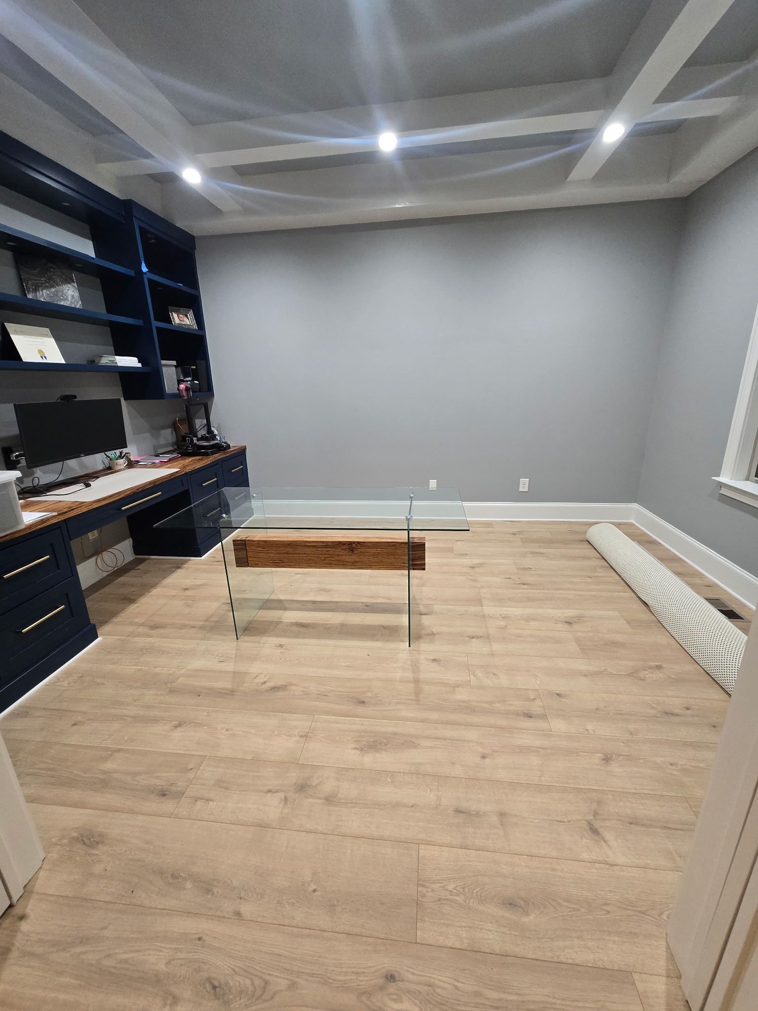 Empty gray home office with built-in shelves, wood floor, recessed ceiling lights, and a small bench in the center