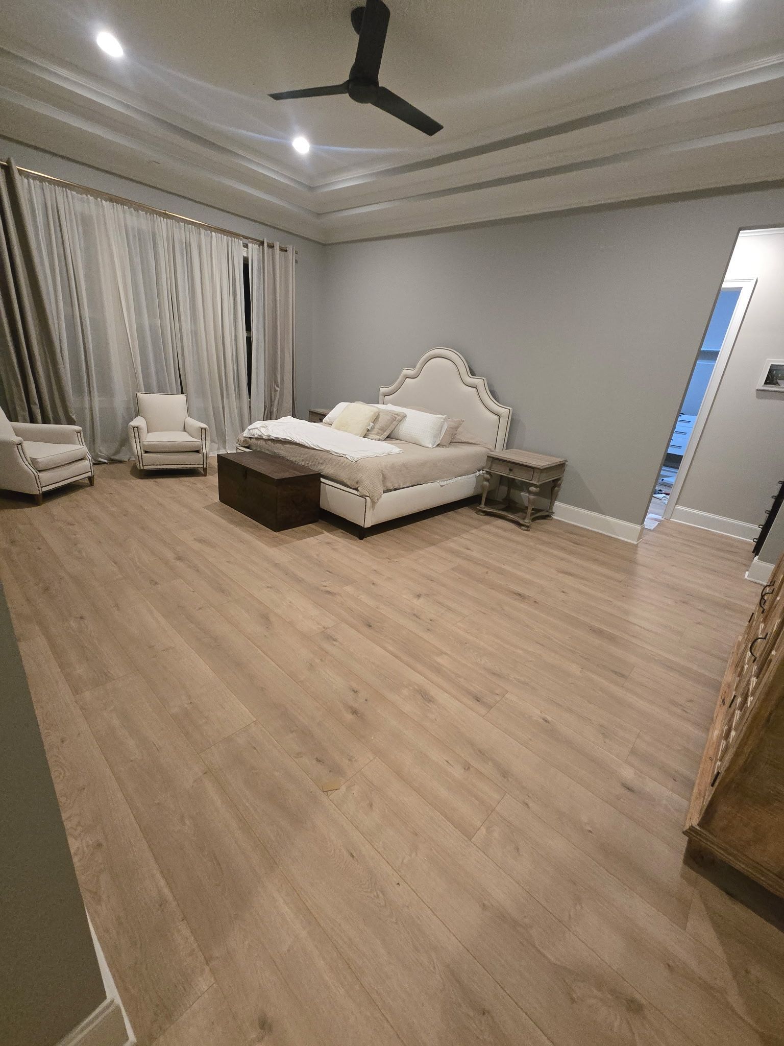 Spacious bedroom with a bed, two armchairs, curtains, ceiling fan, and light wood flooring.