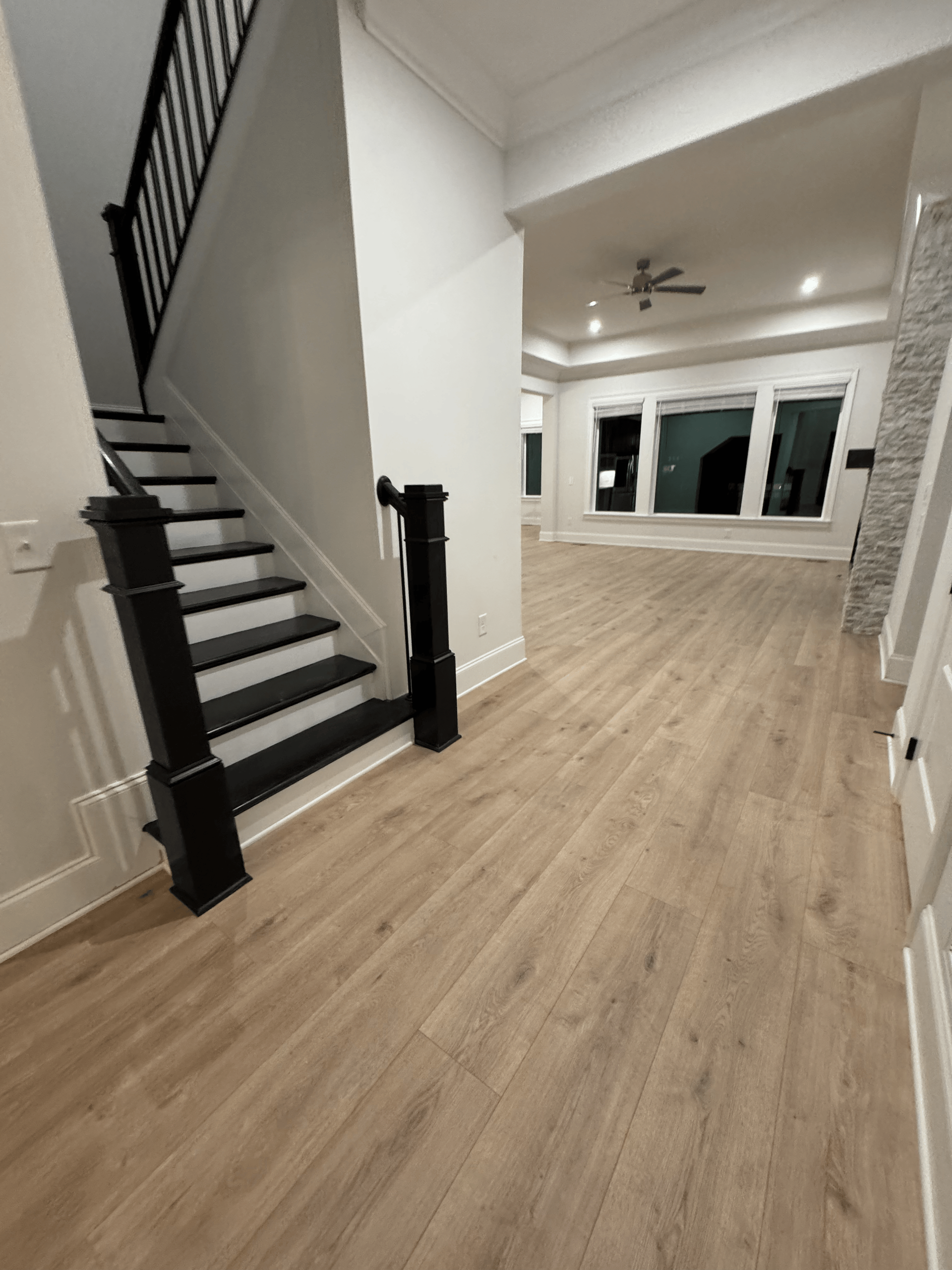 Empty modern living room with light wood floors, black staircase, and large windows.