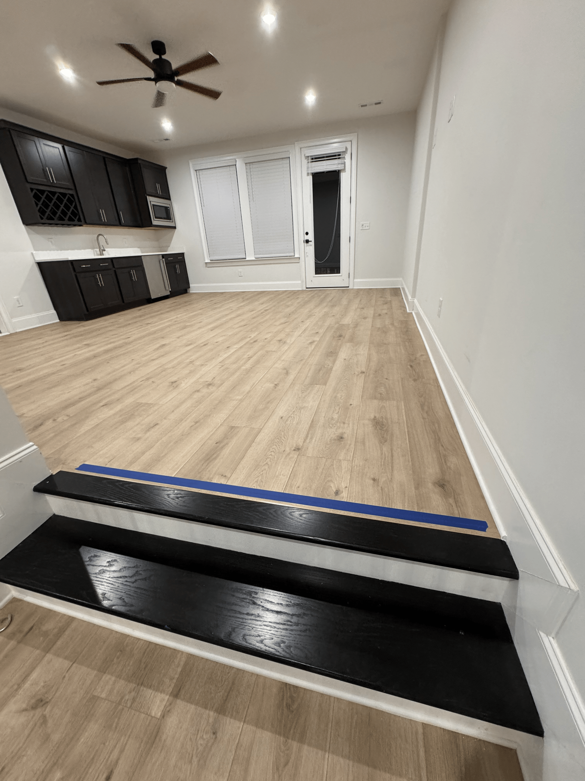 Empty modern room with light wood floors, white walls, black cabinets, ceiling fan, and a small step down at the entrance