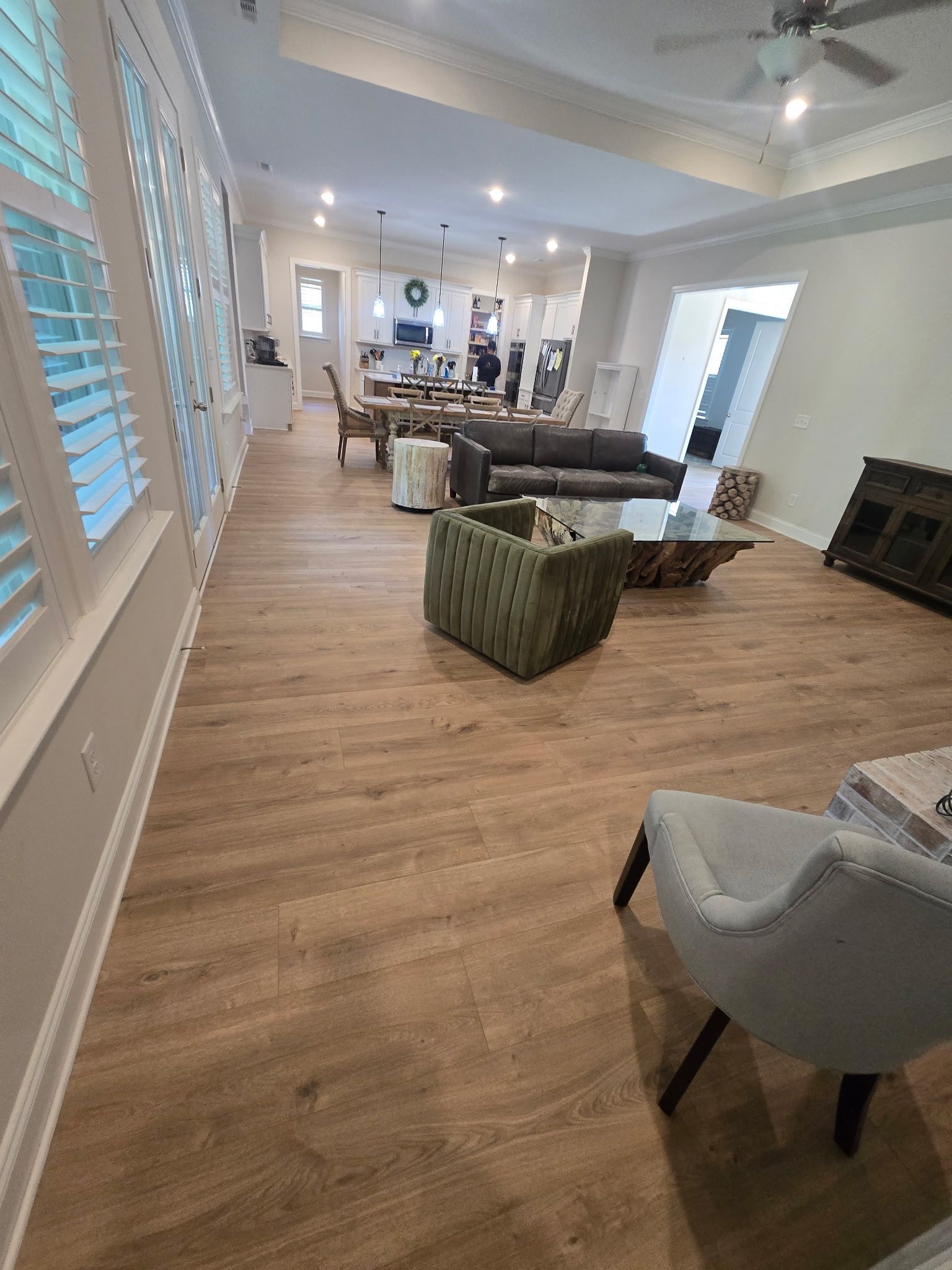 Bright open living room with wood floors, white shutters, gray chairs, green ottoman, and a sofa near the back.