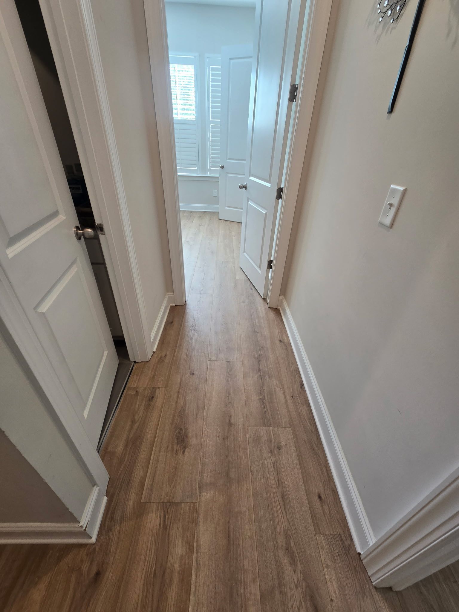 Narrow hallway with wood floors and white walls leading to a bright room at the end.