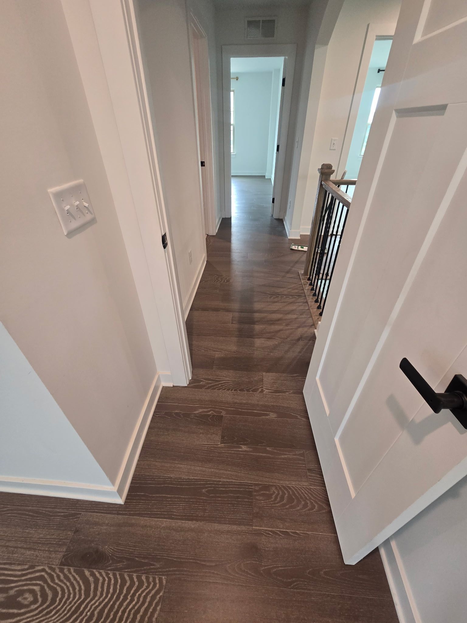 Hallway with dark wood floor, white walls, open door, and stair railing leading to a bright room.