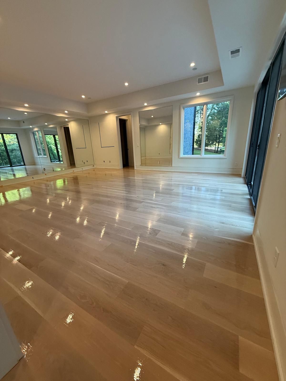 Empty modern living room with polished wood floors, recessed lights, and large windows overlooking greenery