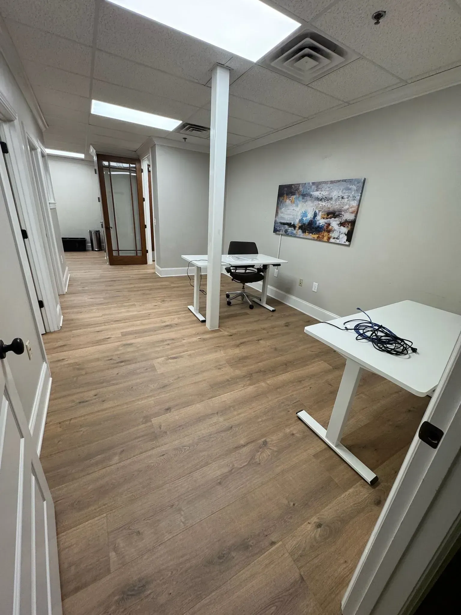 Empty office with white desks, wood flooring, and a wall poster under bright ceiling lights.