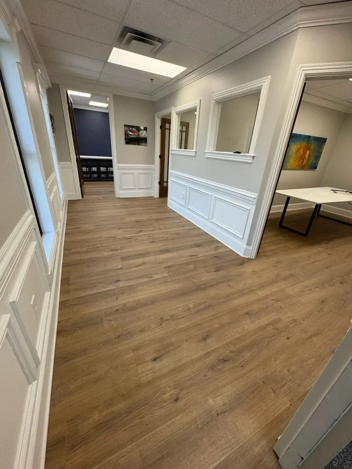 Empty office hallway with white wainscoting, wood floors, and a small table along the right wall.
