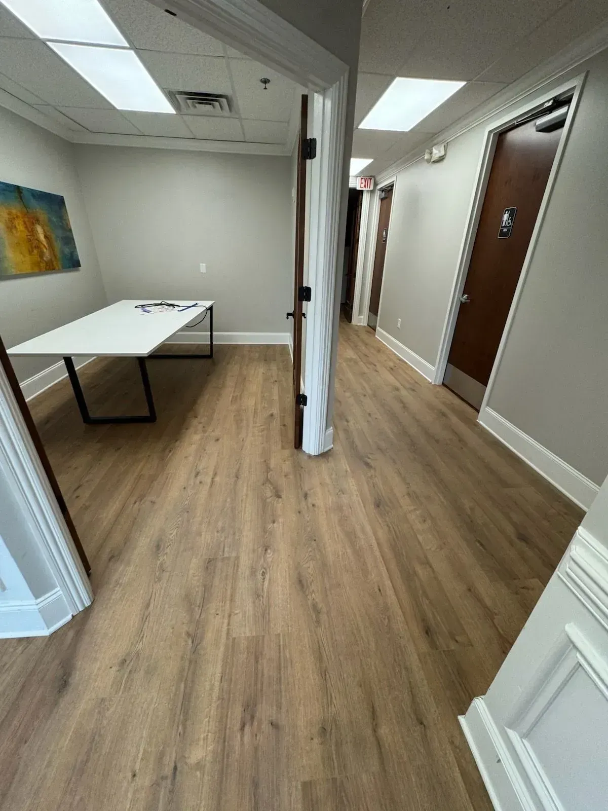 Empty office hallway with wood floors, white walls, a table, and brown doors
