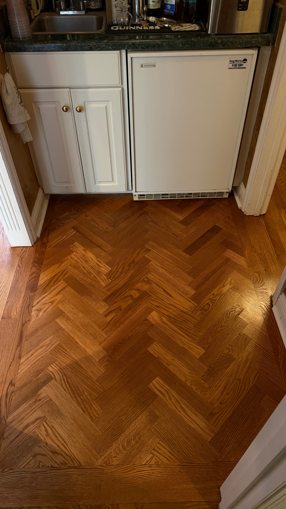 White freezer and refrigerator beneath a cabinet, with a polished herringbone wood floor in front.