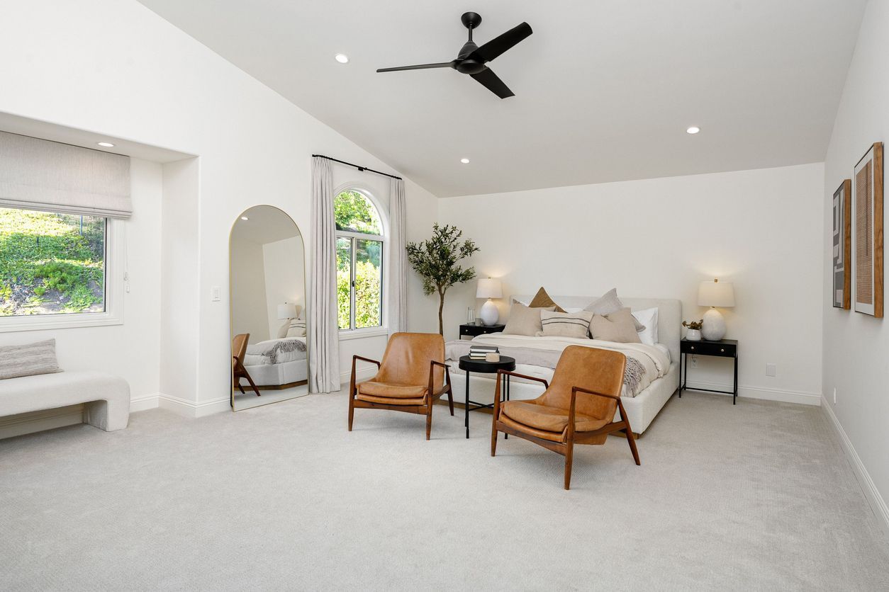 Bright minimalist bedroom with white walls, beige carpet, wooden chairs, and a bed with soft neutral bedding