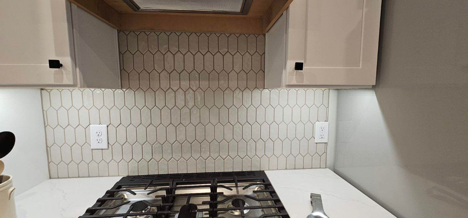 Modern kitchen backsplash with white hex tile, stove, cabinets, and under-cabinet lighting.