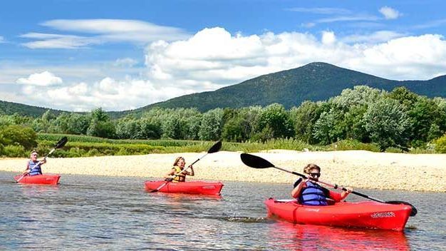 Three people kayaking in red kayaks on a river with a mountain and blue sky in the background