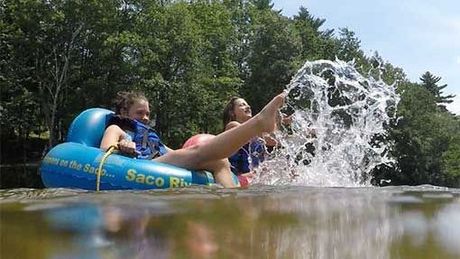 Two people ride a blue tube on a lake, splashing water near a wooded shore.