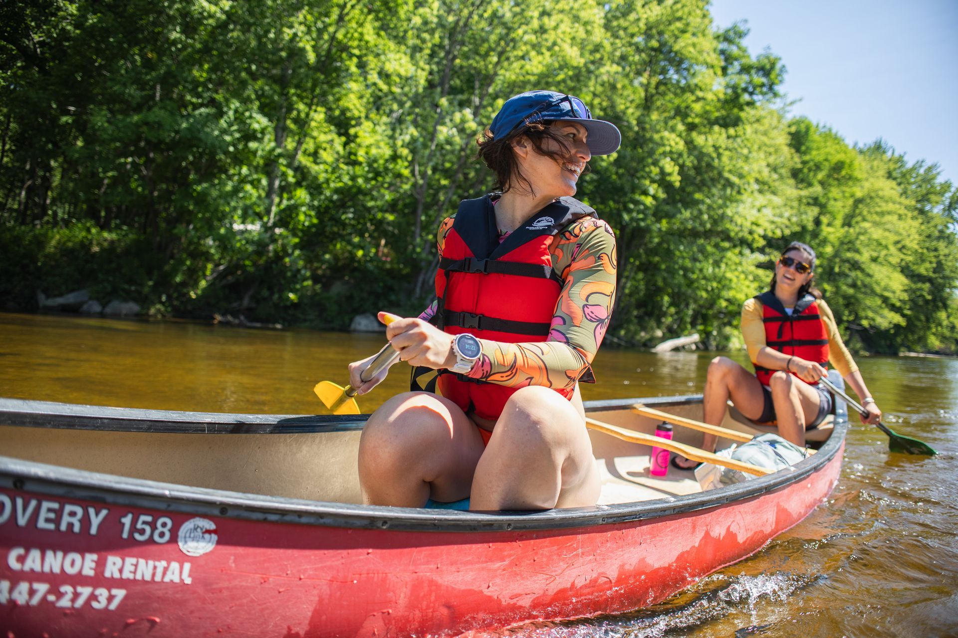People canoeing on a river, one in a red canoe wearing a life vest, with green trees along the bank