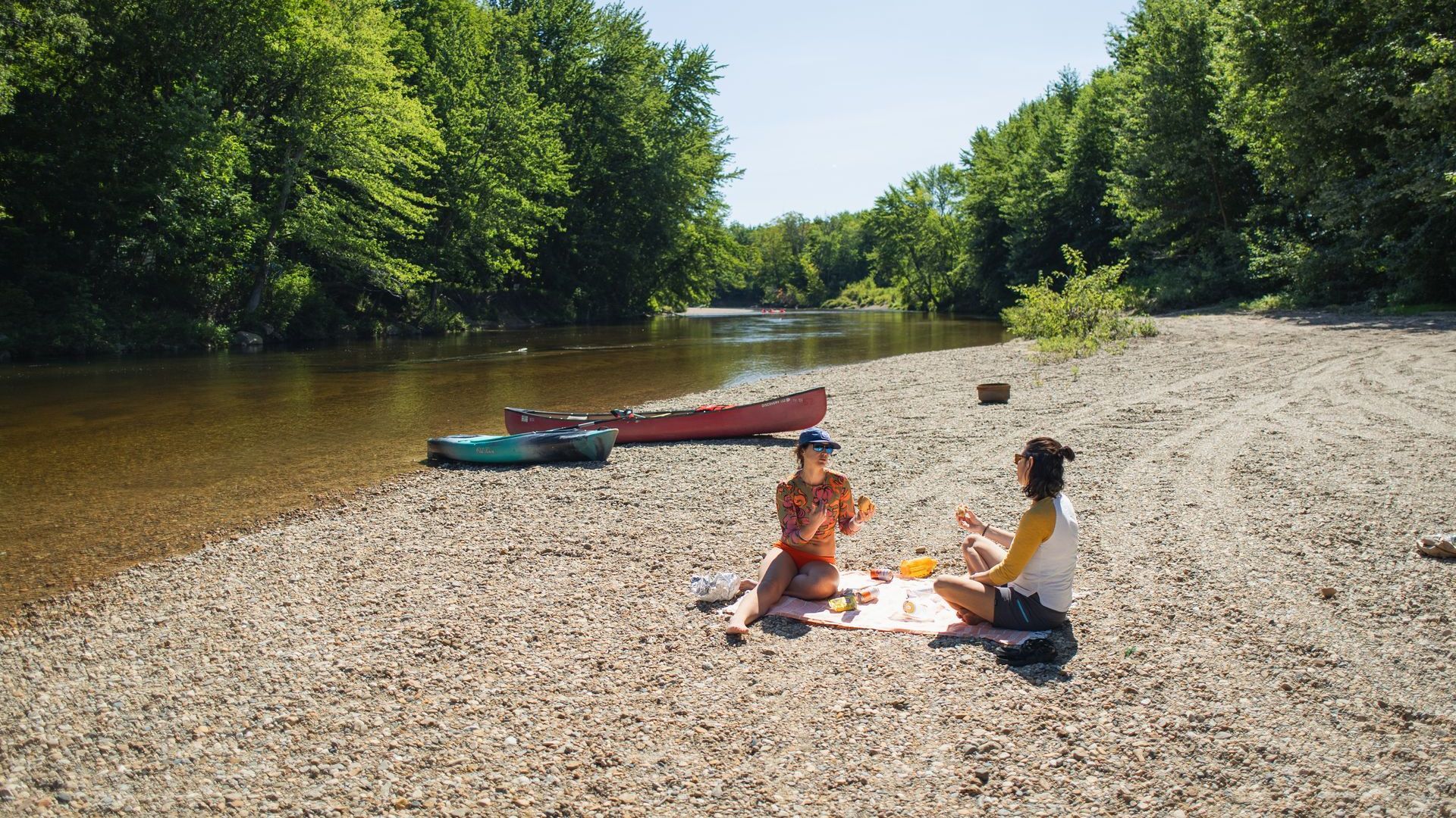Two people sit on a rocky riverbank beside a canoe on a calm, tree-lined river.