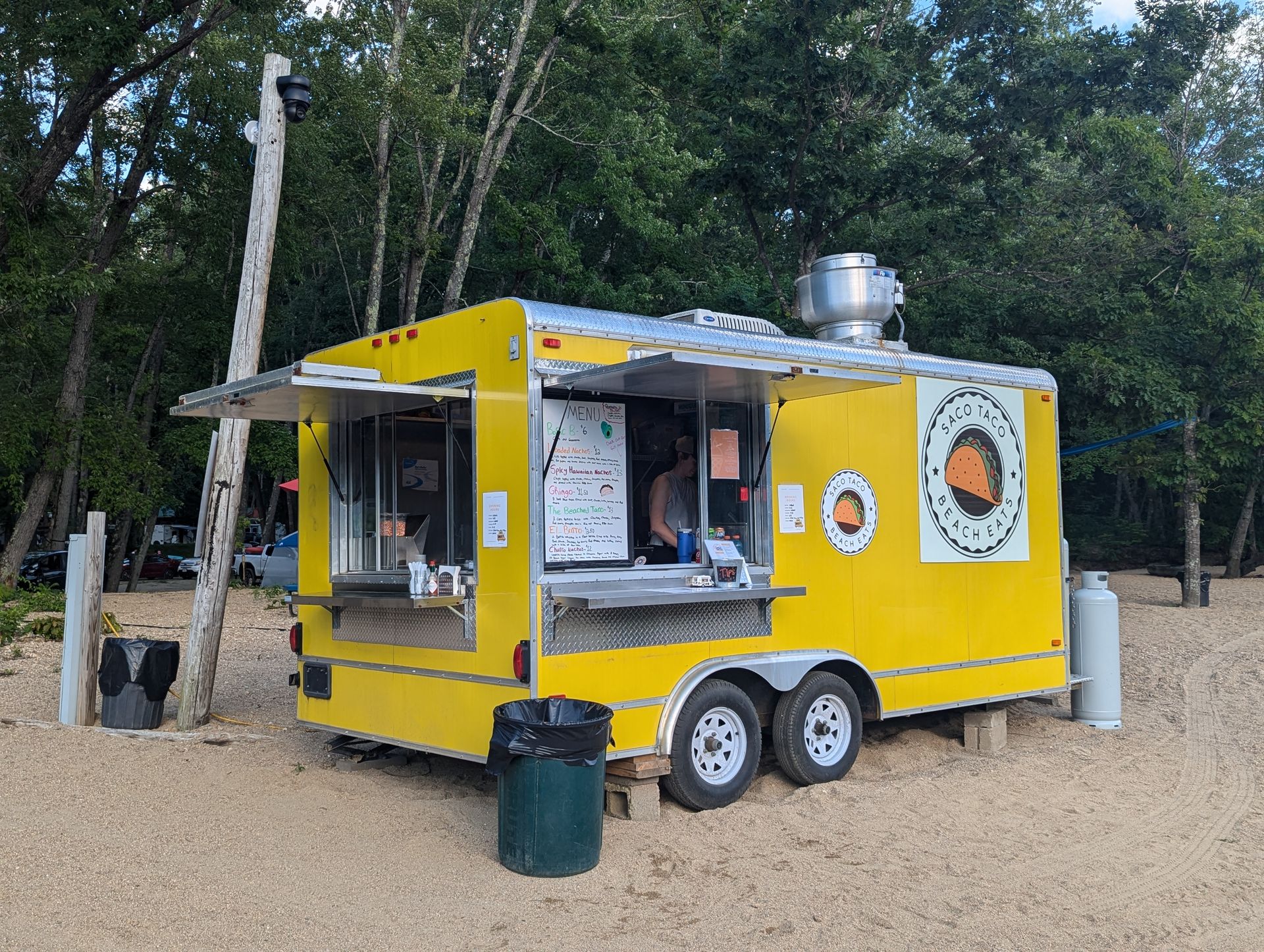 Yellow food trailer with open serving window and round porthole-like windows in a wooded area