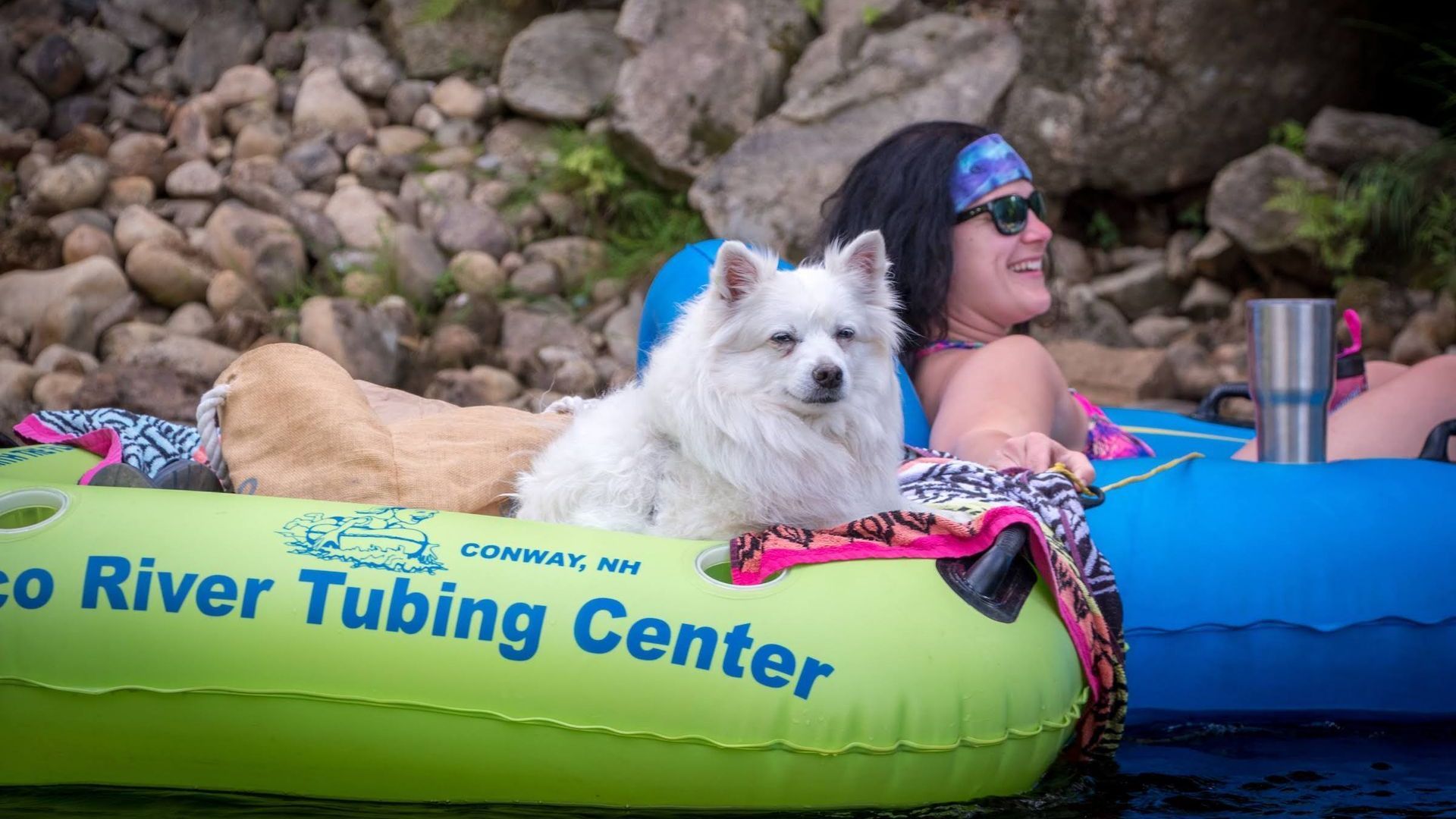 White dog on green river tube with woman in sunglasses floating on water near rocky shore