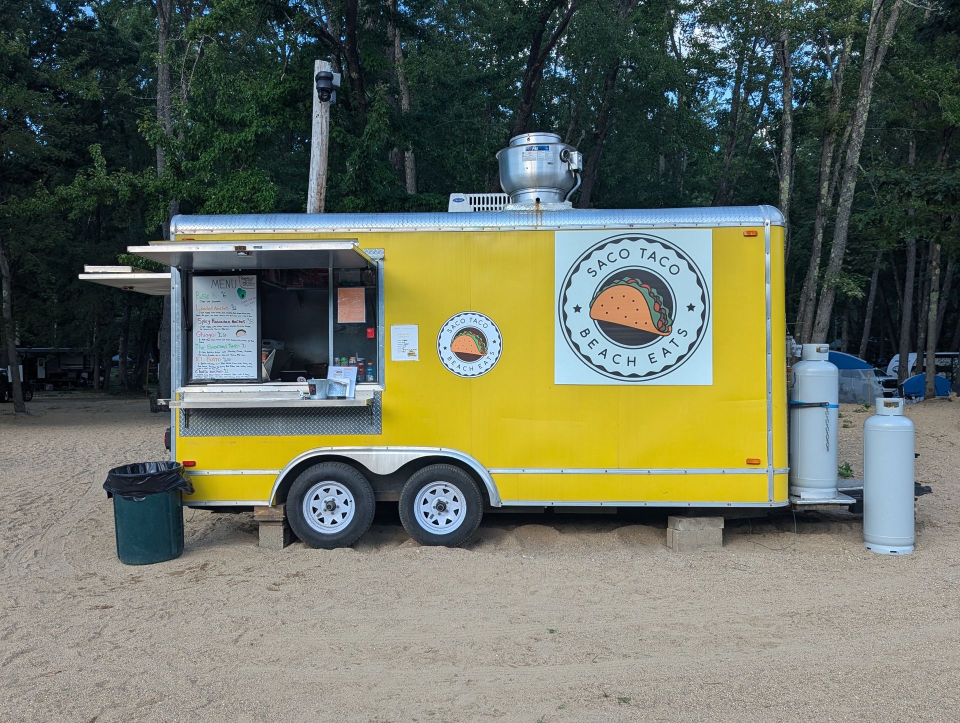 Yellow food truck with serving window, circular logo, and propane tanks parked in a wooded area