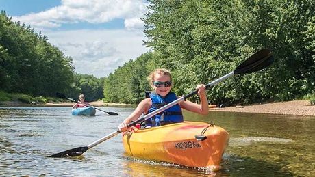 Child kayaking in a yellow kayak on a river, smiling and paddling, with another kayaker behind.