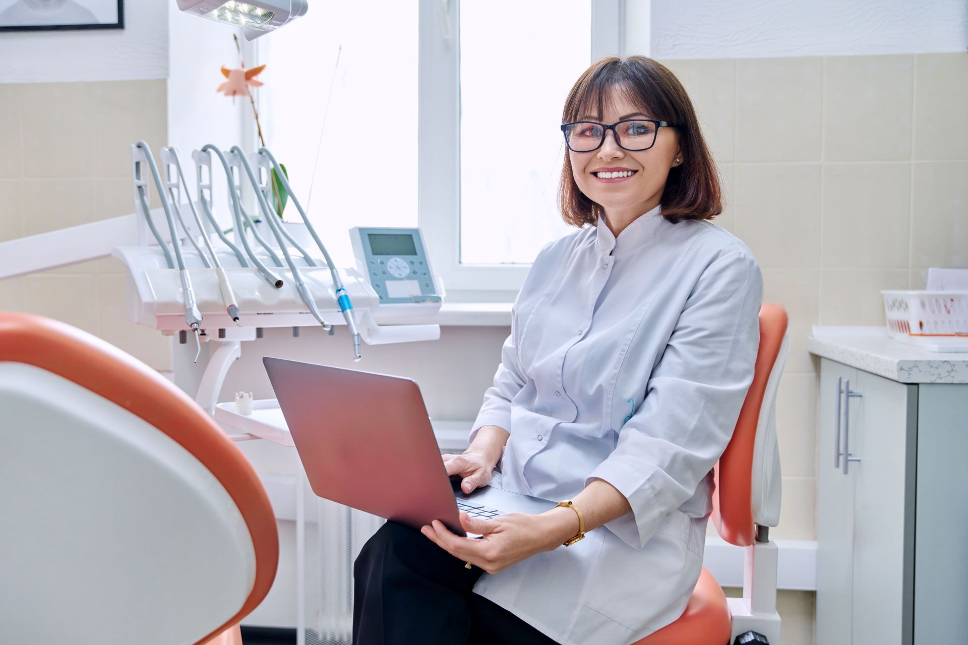 Dentist holding laptop, smiling in office with dental equipment.