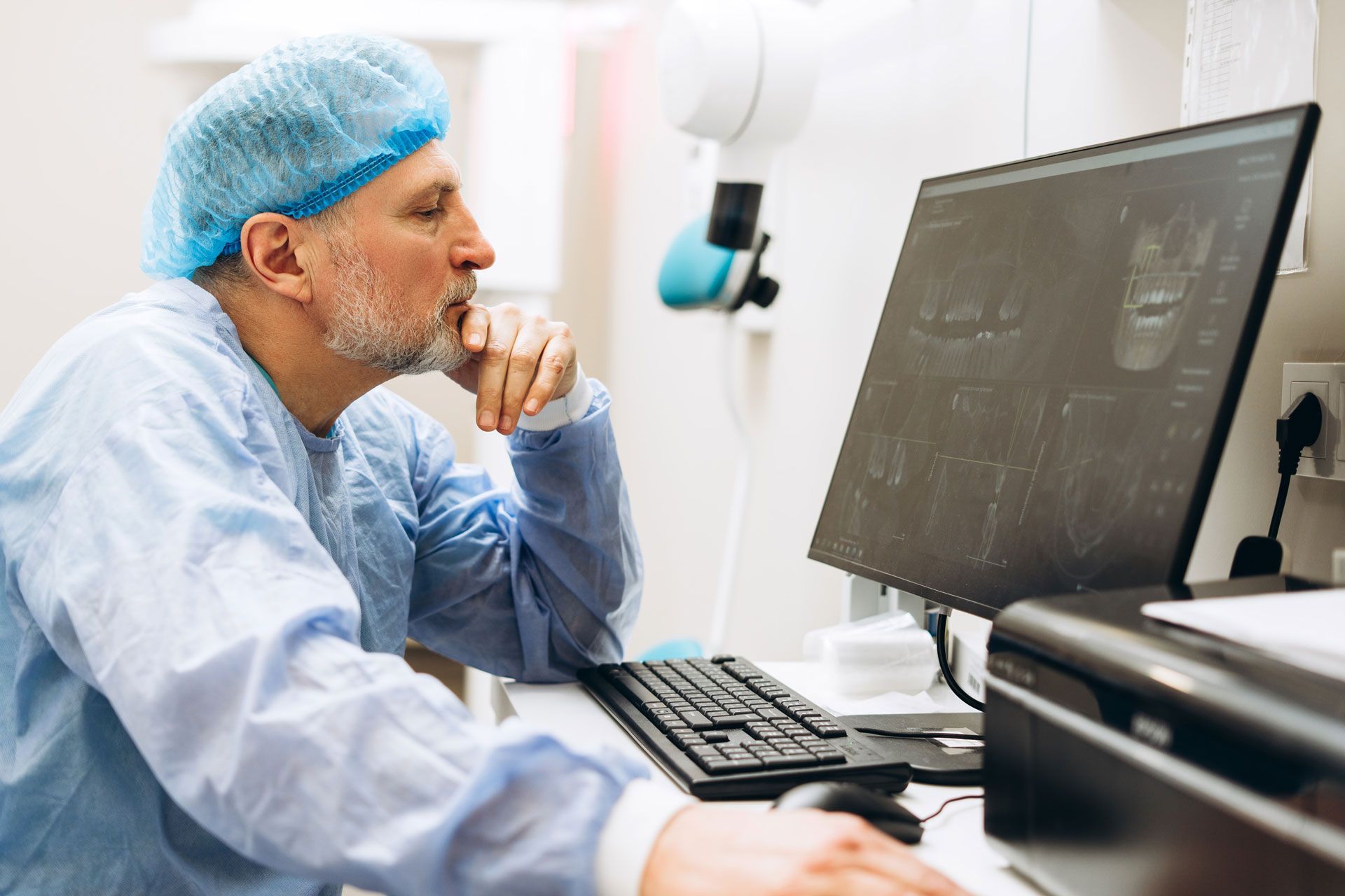 Dentist in blue surgical attire examines dental x-rays on a computer monitor.