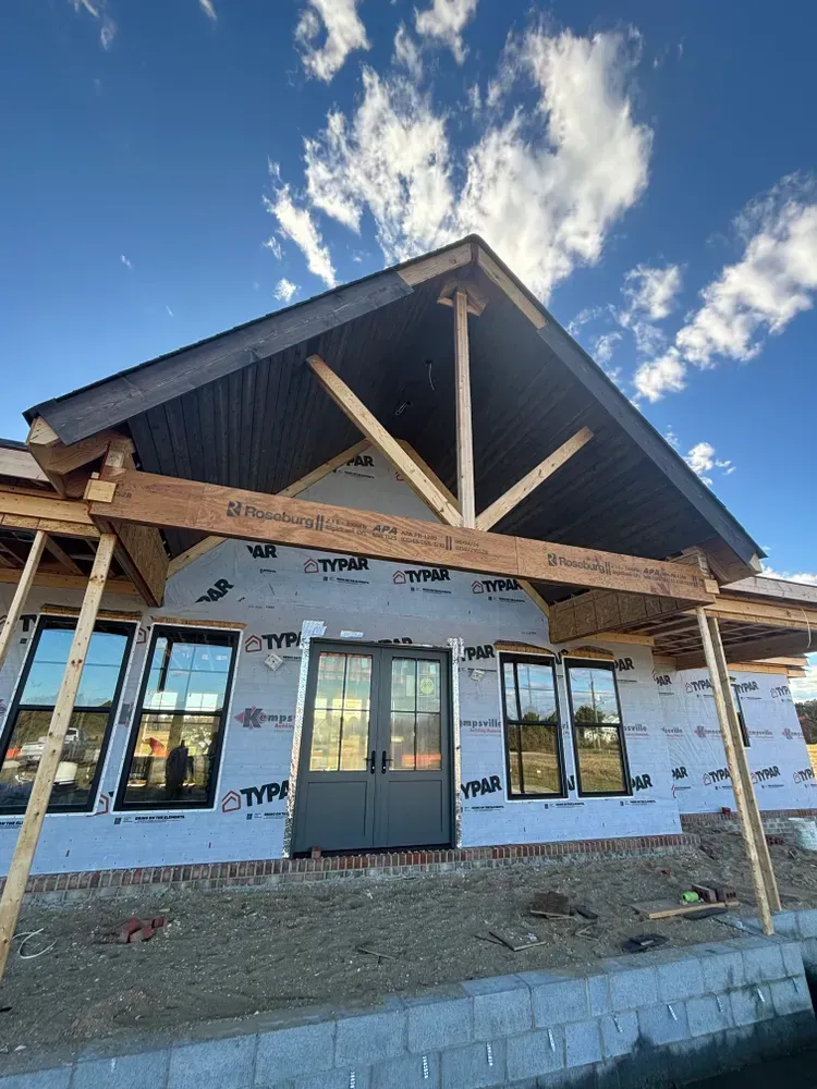 Front view of a house under construction with white wrap, dark roof, and large front windows under a blue sky