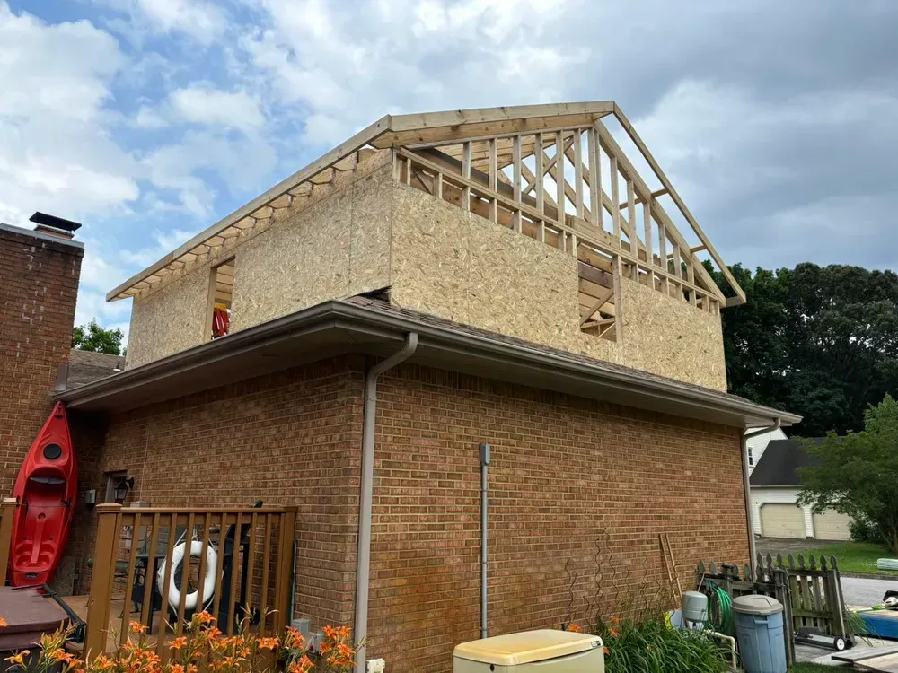 Two-story brick house under construction with exposed wooden roof framing and a red kayak at left.