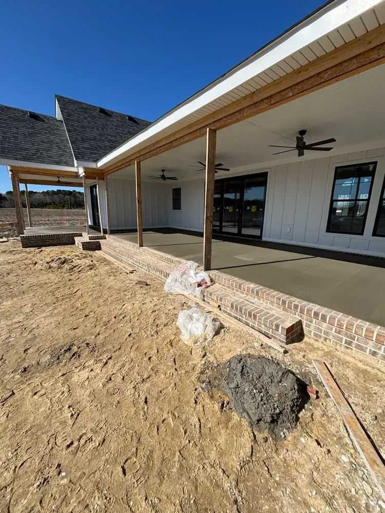 New house exterior with covered patio, brick columns, and unfinished dirt yard under a blue sky
