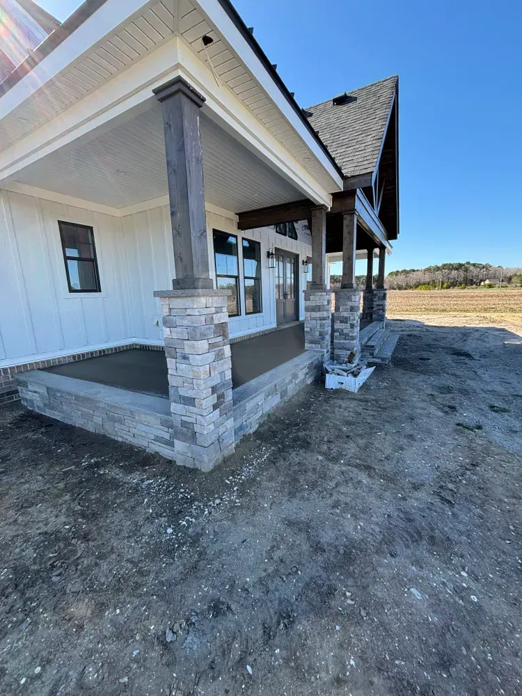 New home exterior with covered porch, stone columns, and unfinished dirt yard under a clear sky